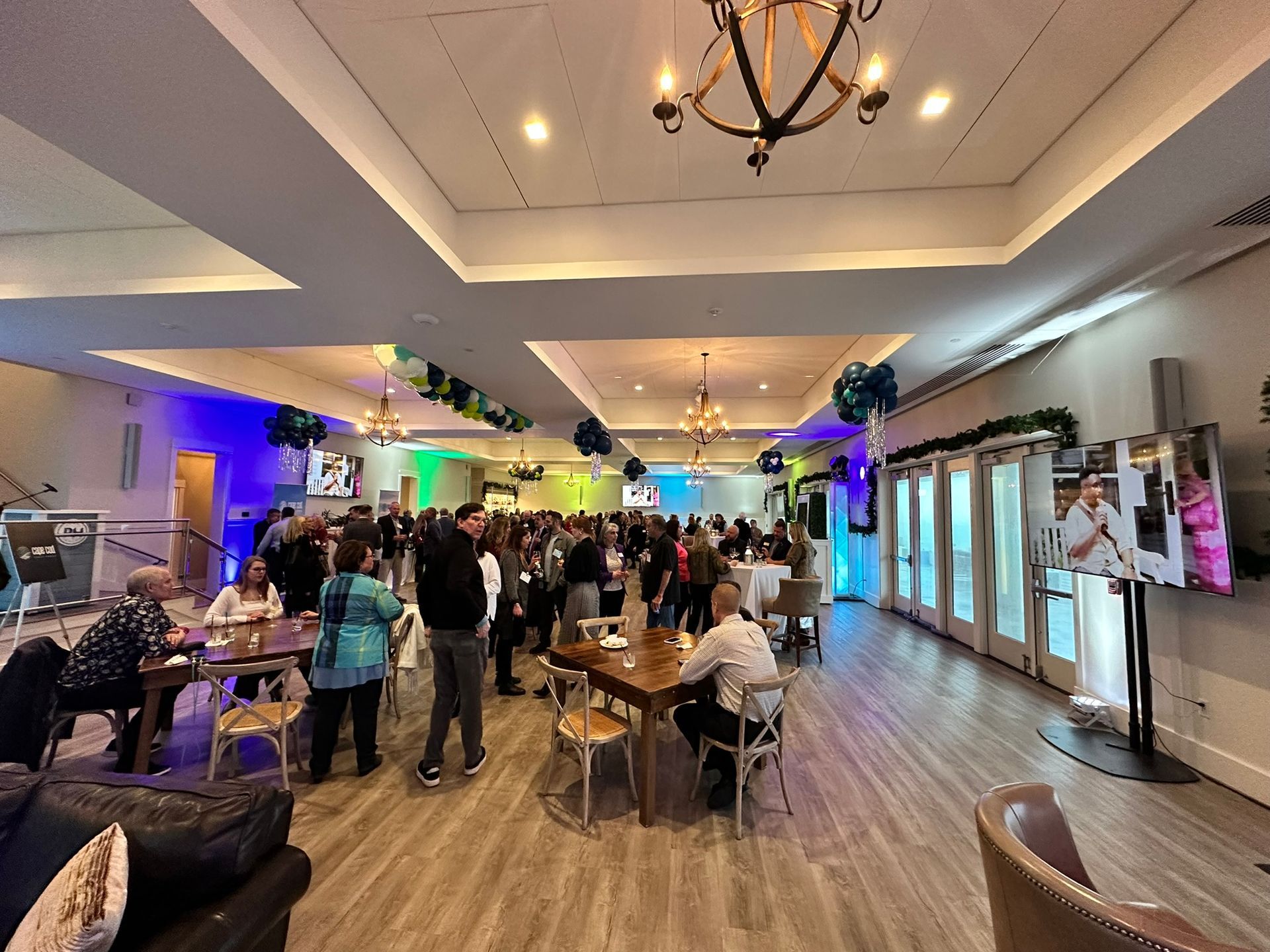 Event in a large room with tables, people mingling, and screens showing a speaker.