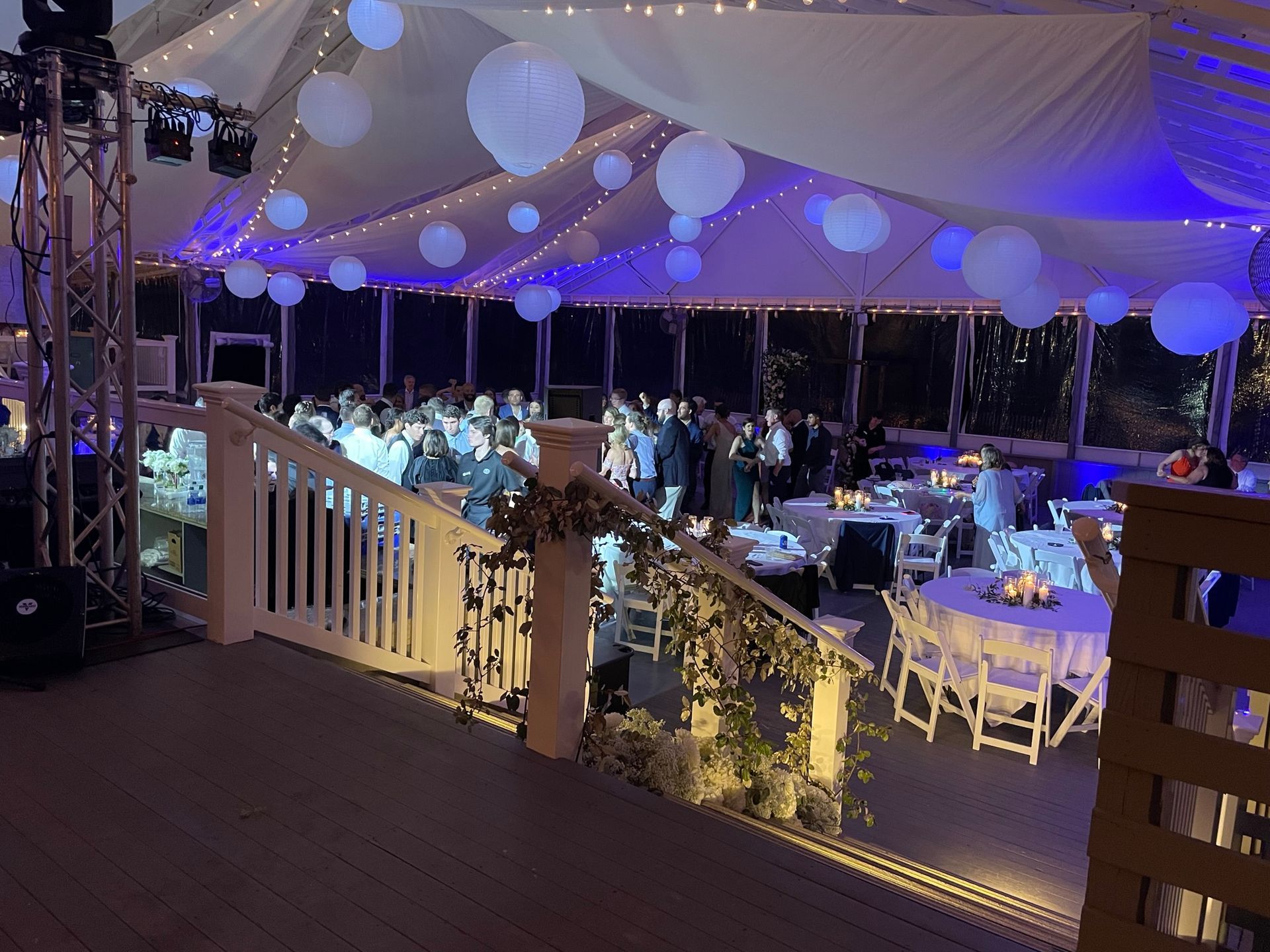Wedding reception inside a tent with white balloons, tables, and dancing guests. Blue and white lighting.