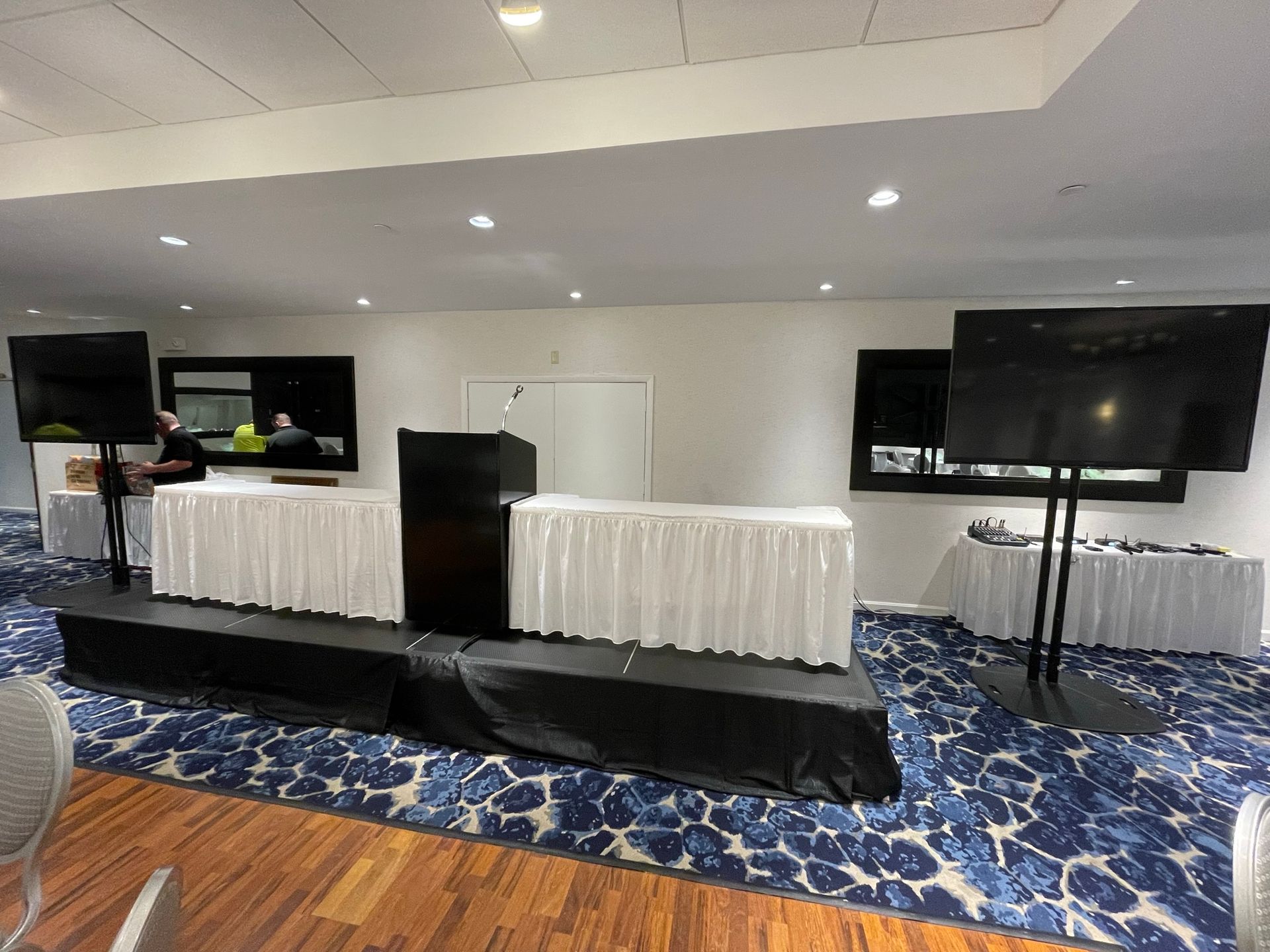 Conference room setup with stage, tables with white skirting, large screens, and blue patterned carpet.