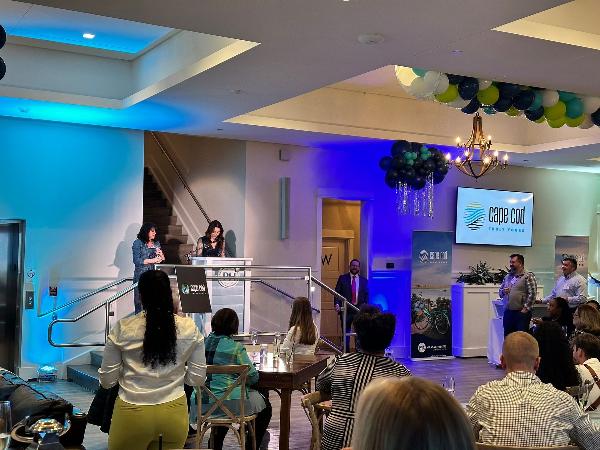 People at an event, two women at a podium. Balloons and a logo on the wall.