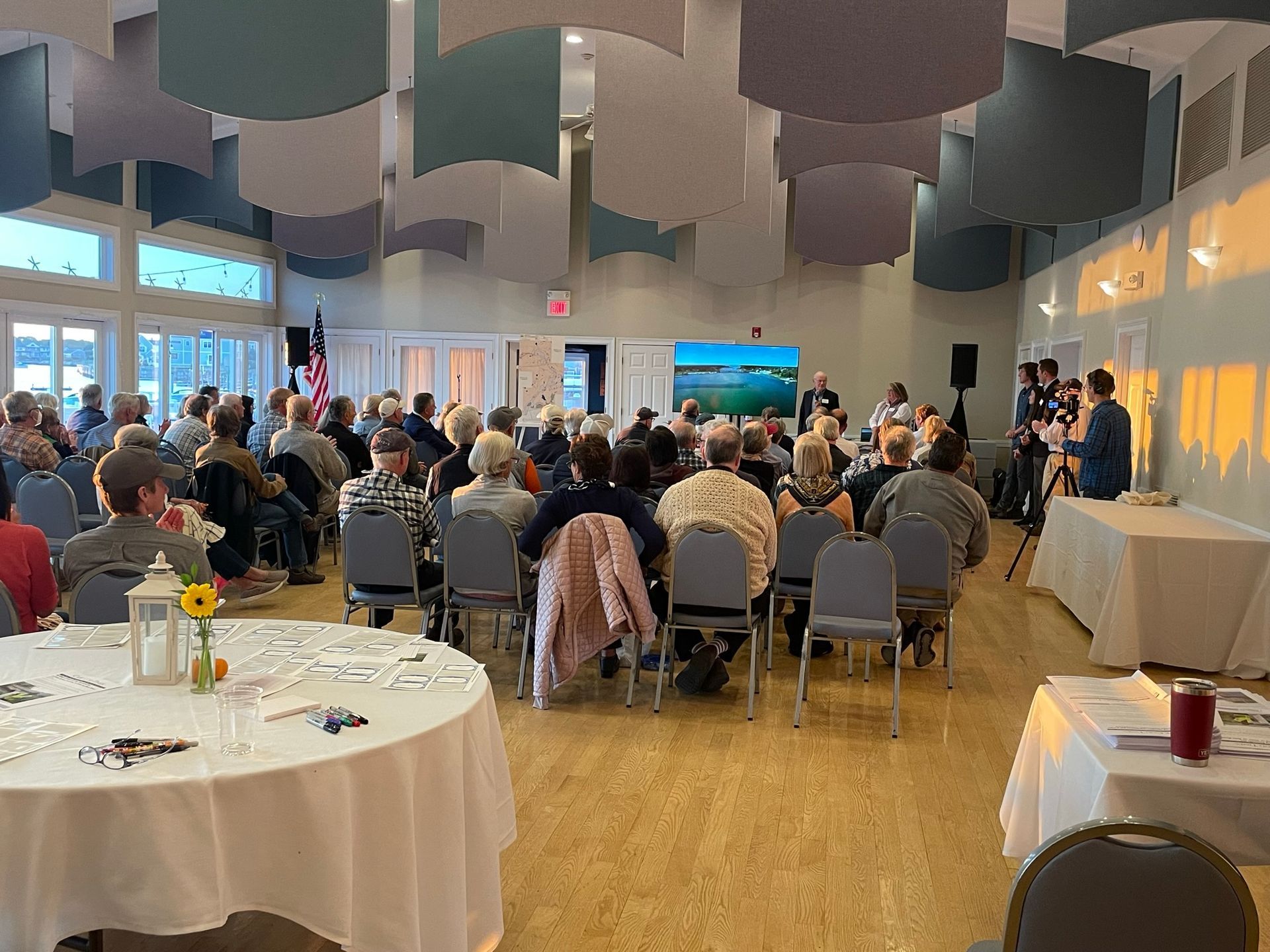 Large room with audience attending a meeting. Speakers at the front near a screen, tables with literature in the foreground.