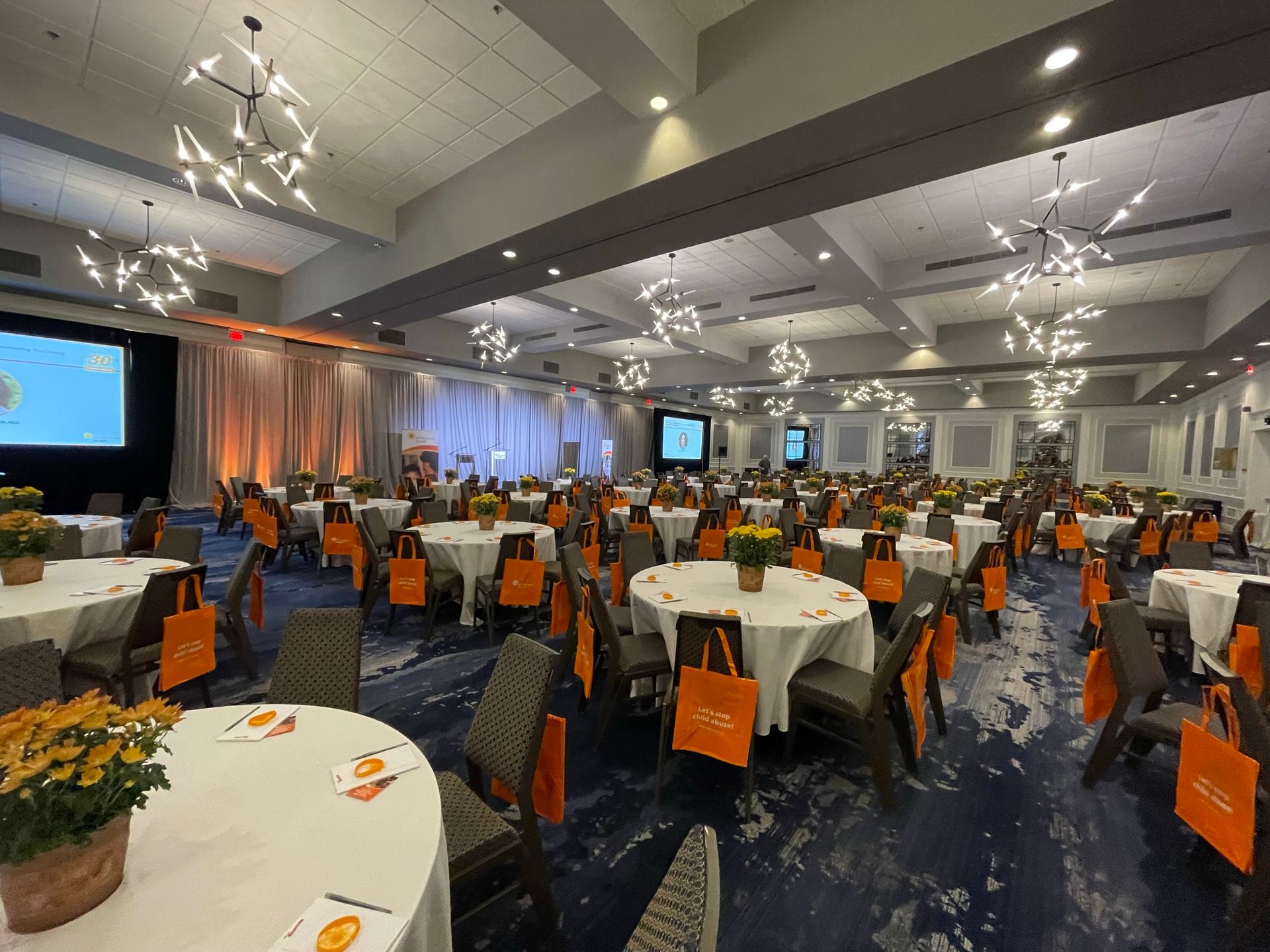 Large conference room set with round tables, chairs, and orange gift bags. Stage with screens and lighting.