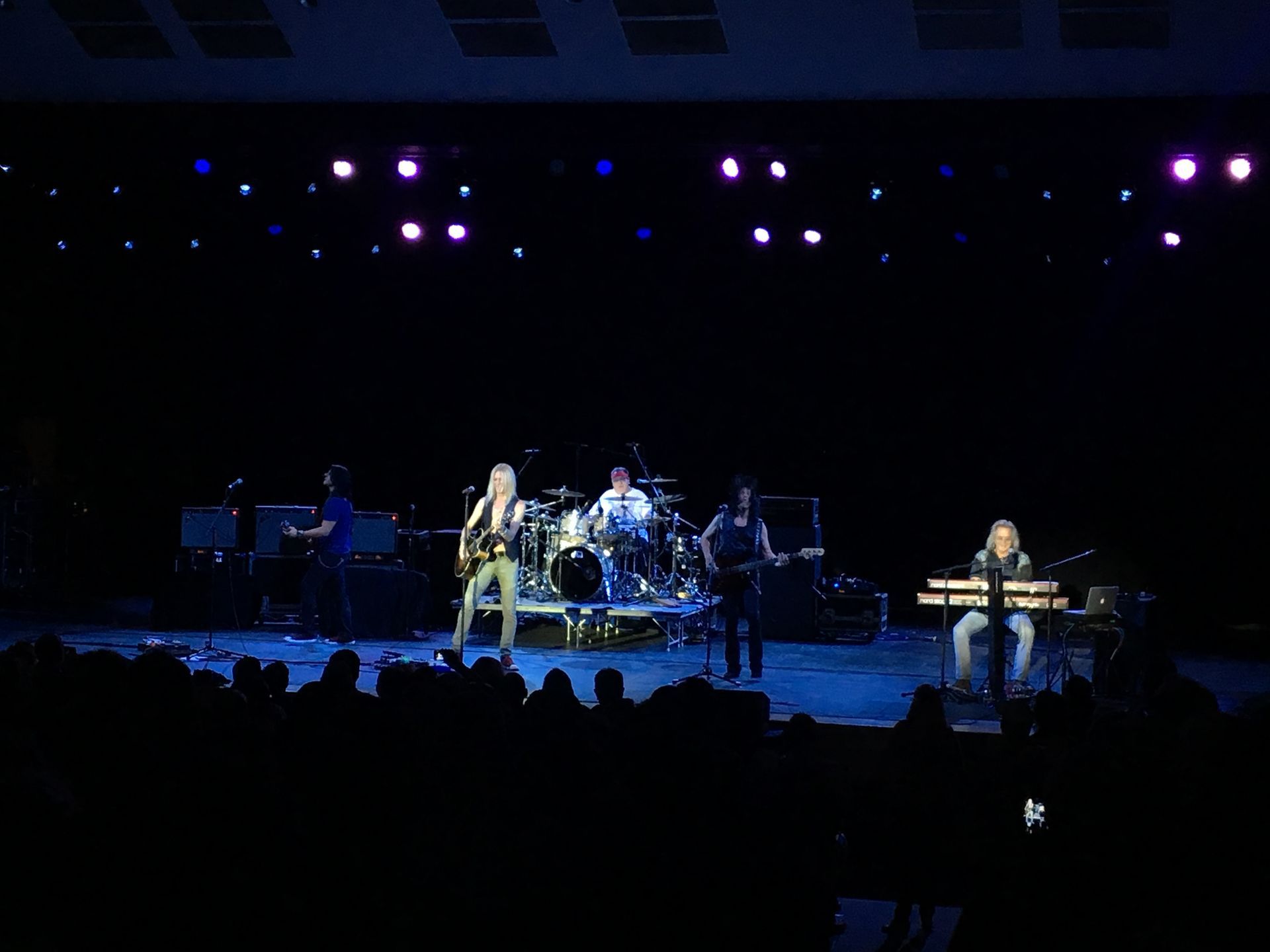 Band performing on stage under stage lights; audience in foreground.