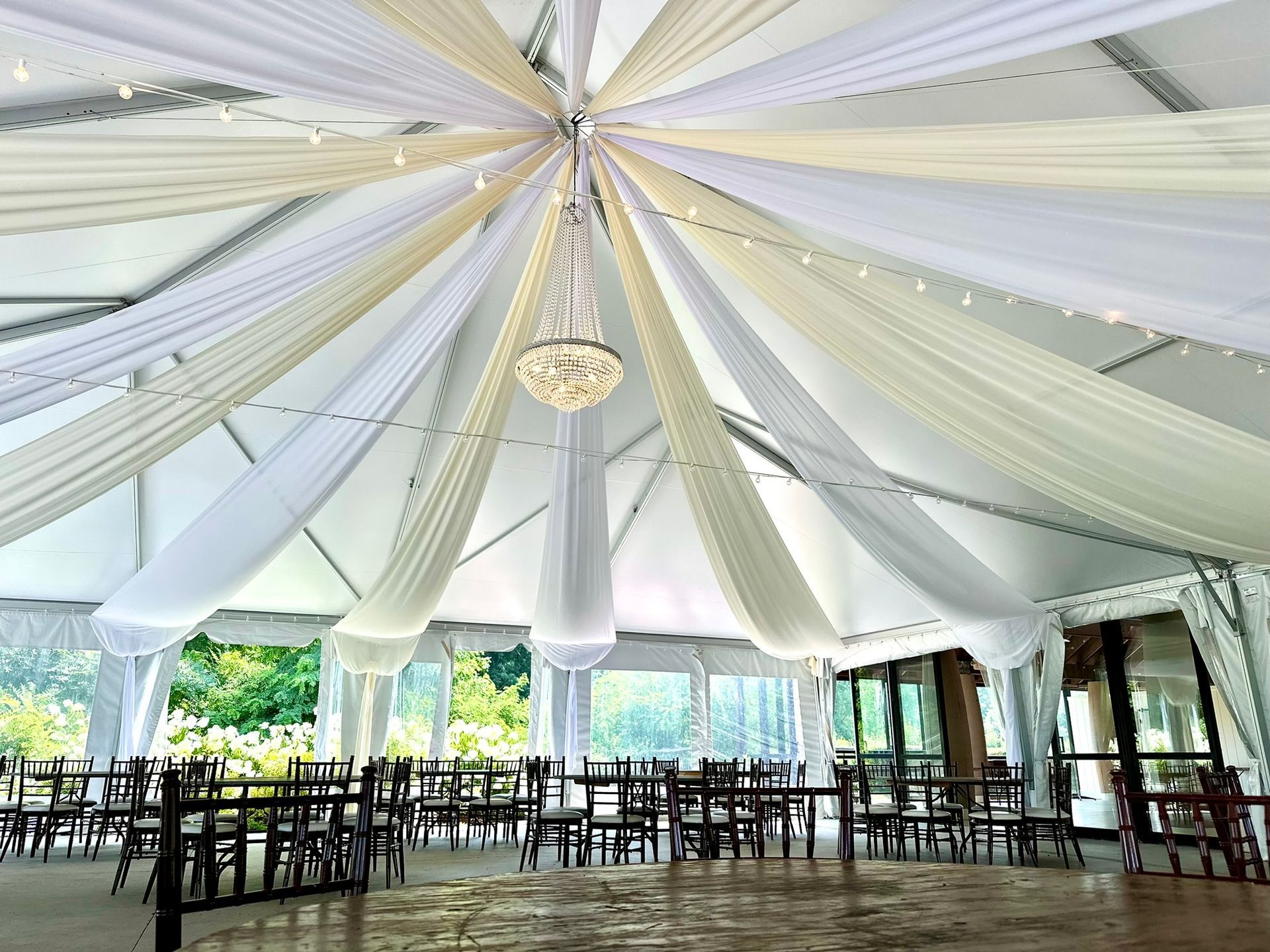 White tent interior decorated with draped fabric and chandelier, tables and chairs set up for an event.