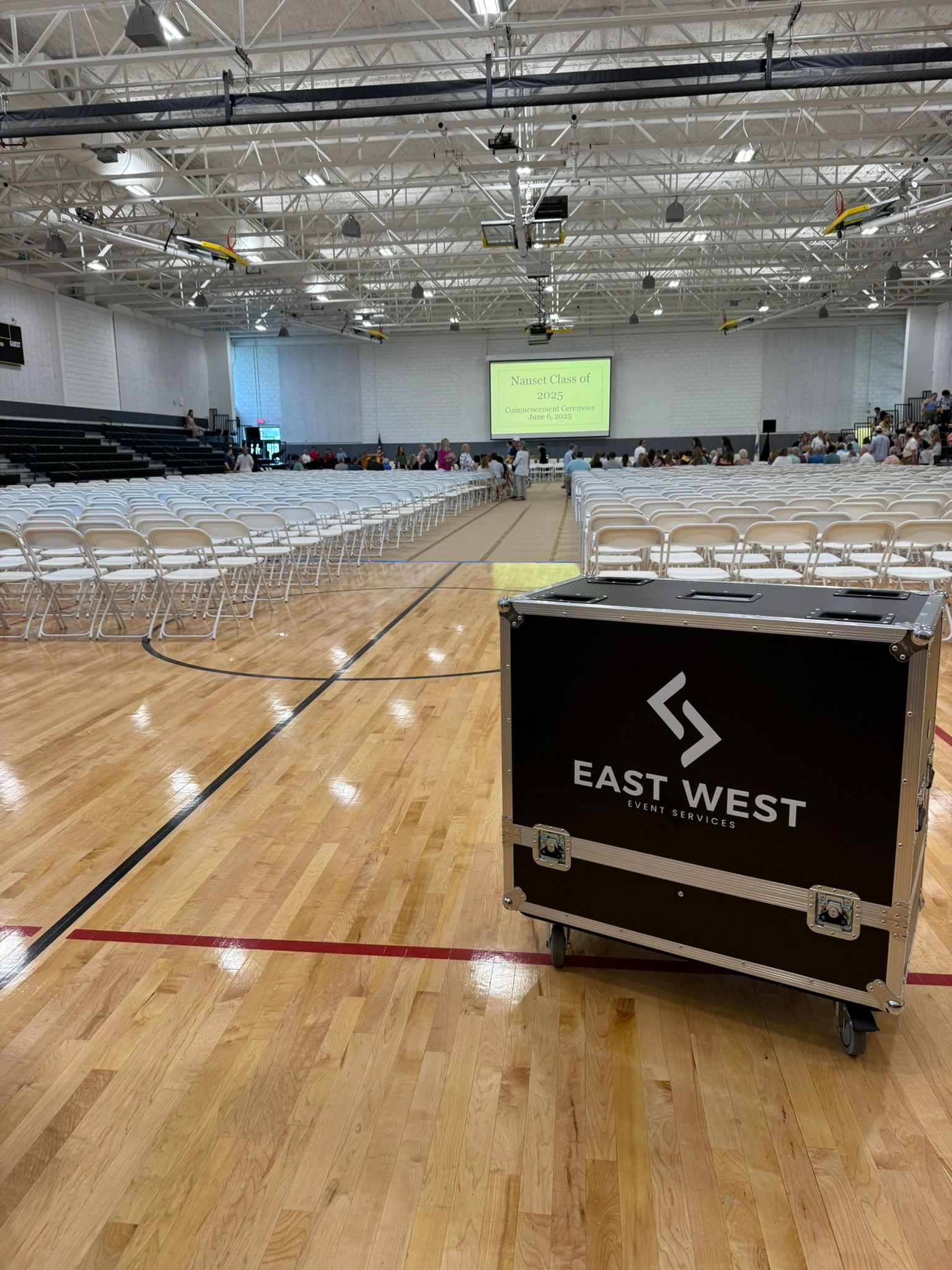 Black equipment case on a gymnasium floor with rows of chairs set up for an event. A screen is visible in the background.