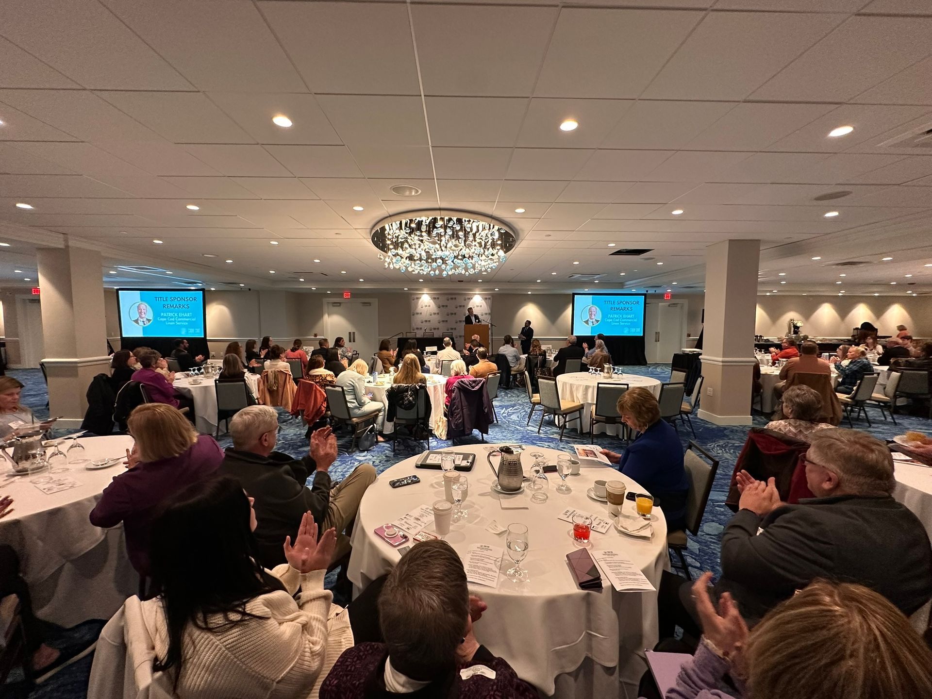 Large ballroom with people at tables, clapping. Two screens display text; a speaker at a podium.