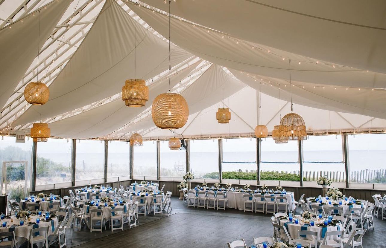 Wedding reception tent overlooking the ocean, decorated with round woven lamps, tables with blue accents.