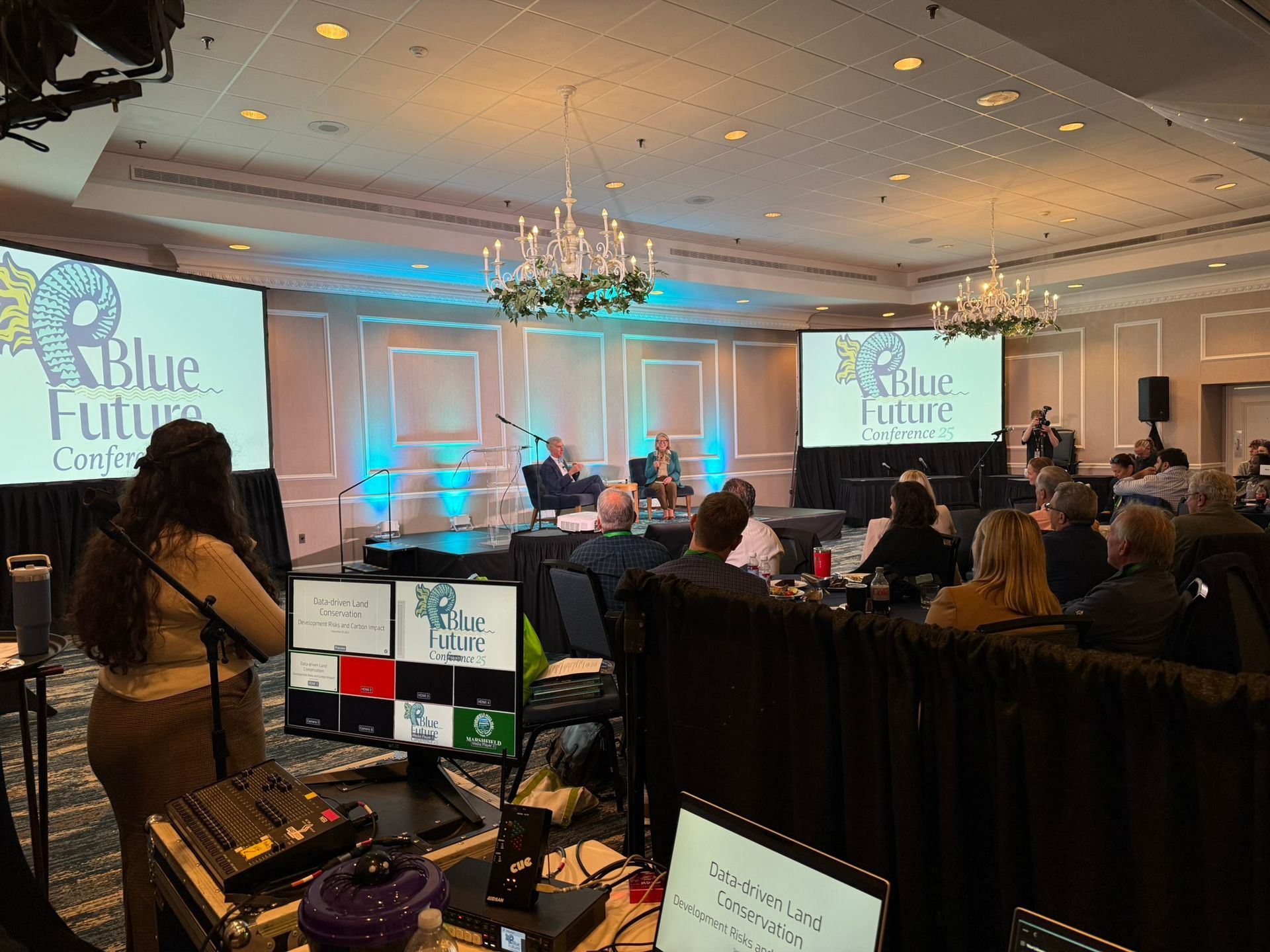 Conference event: Speakers on stage, audience seated. Blue Future logo on screens. Production setup visible.