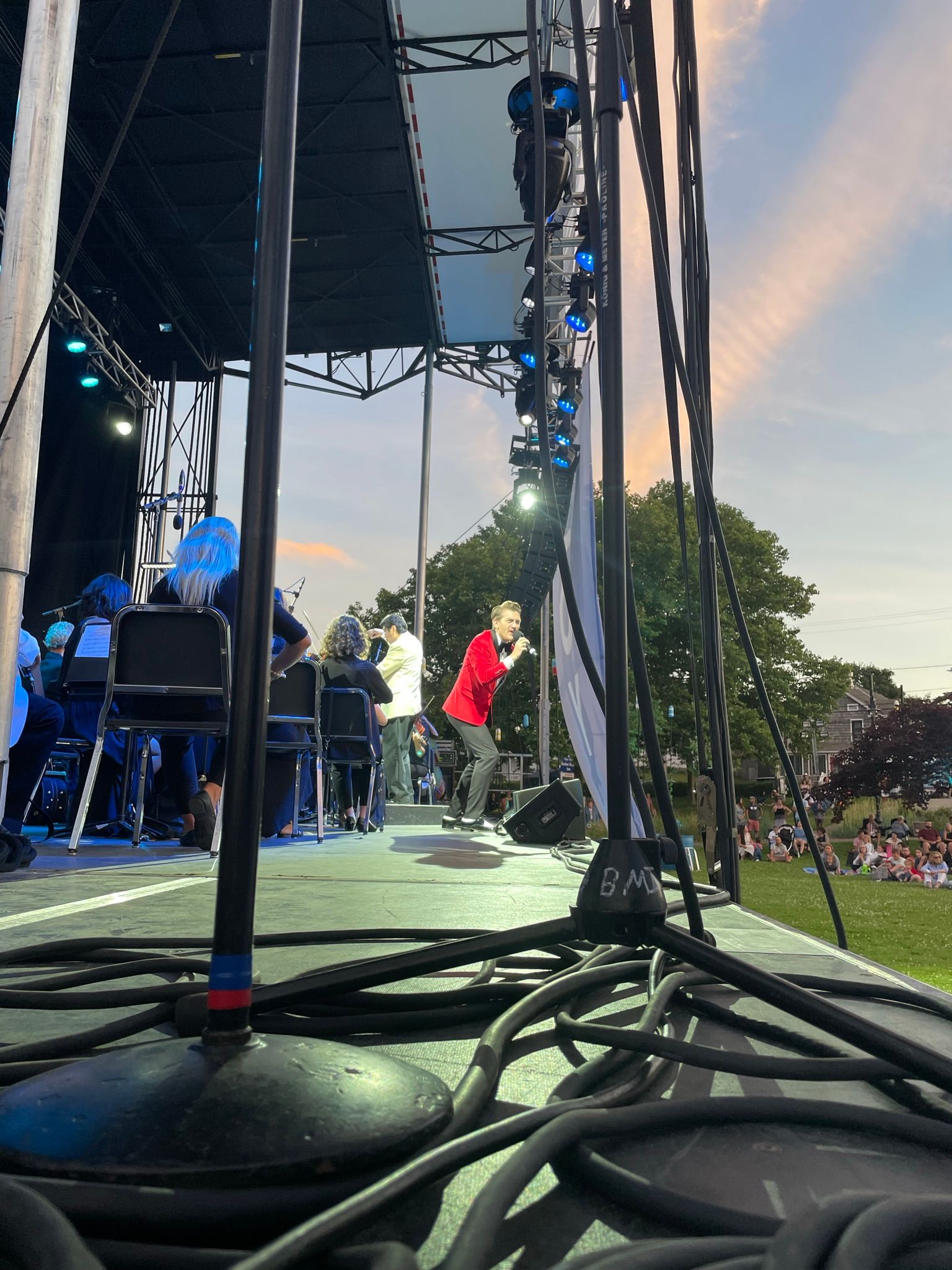 A man in a red jacket performs on stage with an orchestra, cables in foreground.