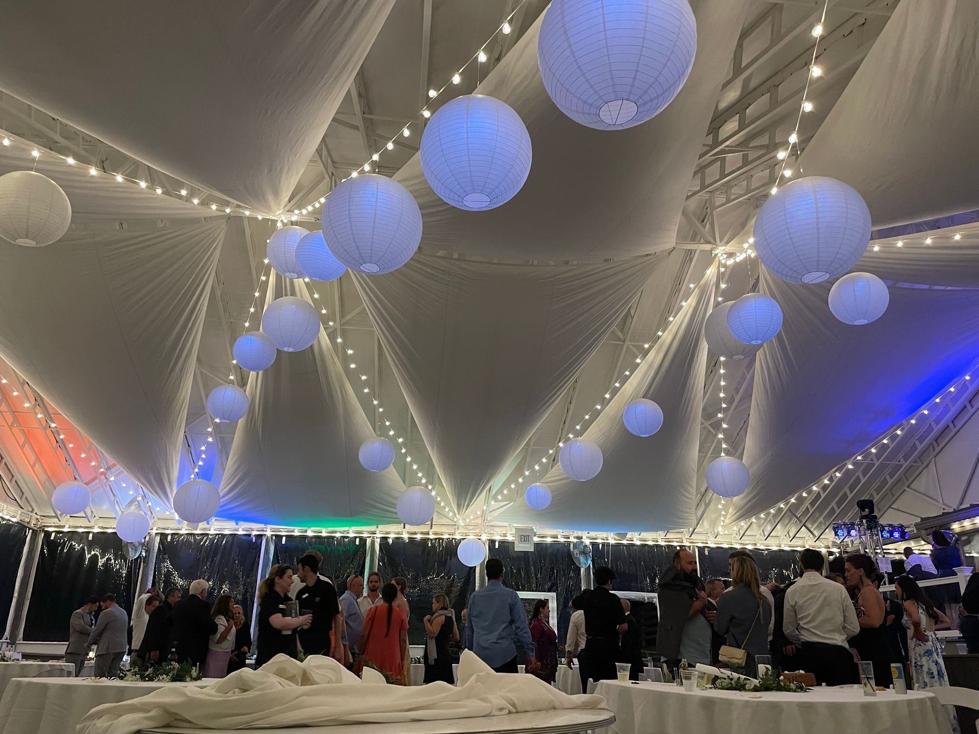 Wedding reception inside a tent, with string lights and white paper lanterns. Guests socialize at tables.