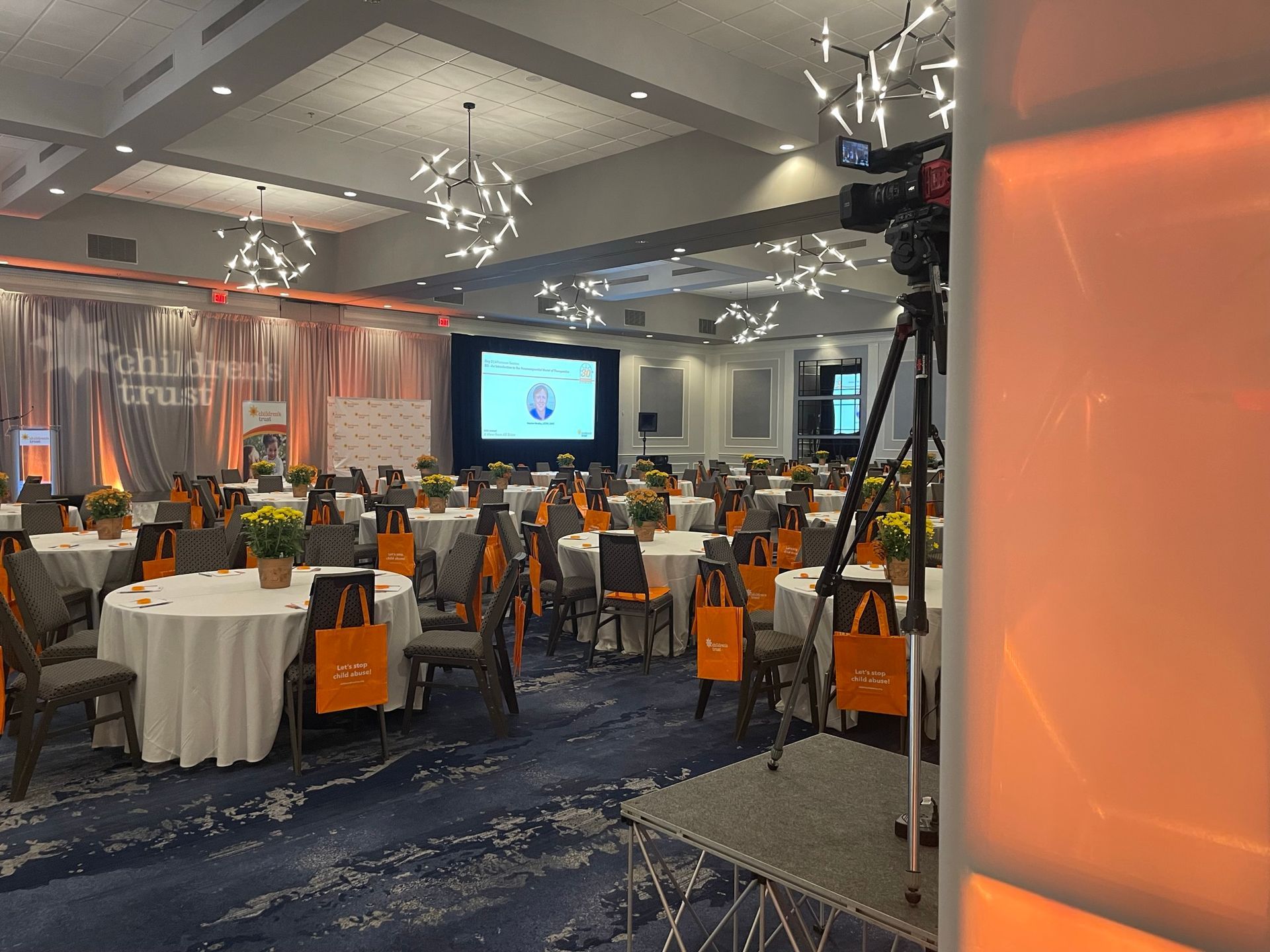 Empty event hall with round tables, orange accents, and a video camera set up.