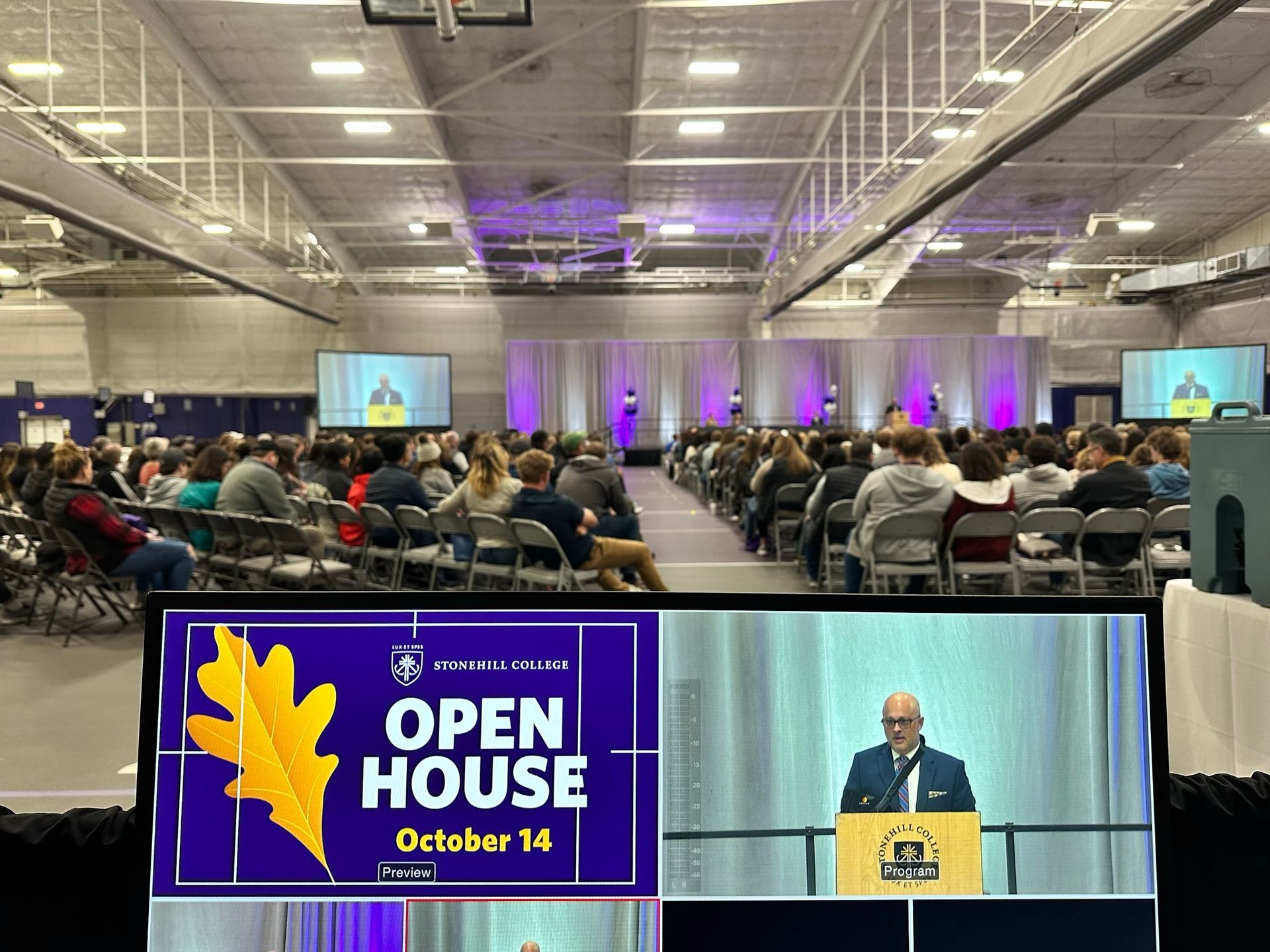 Large open house event in a gymnasium with a speaker at the podium.