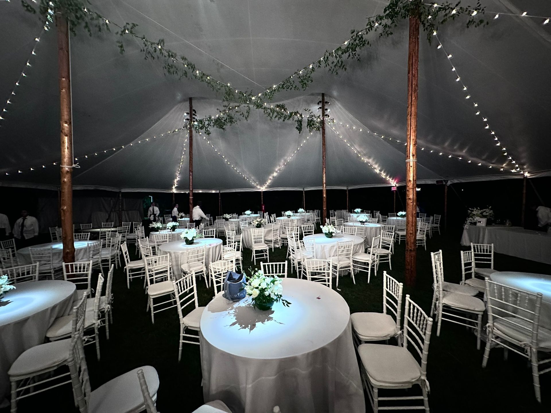 Wedding reception tent with white tables and chairs, strung with lights, and greenery.