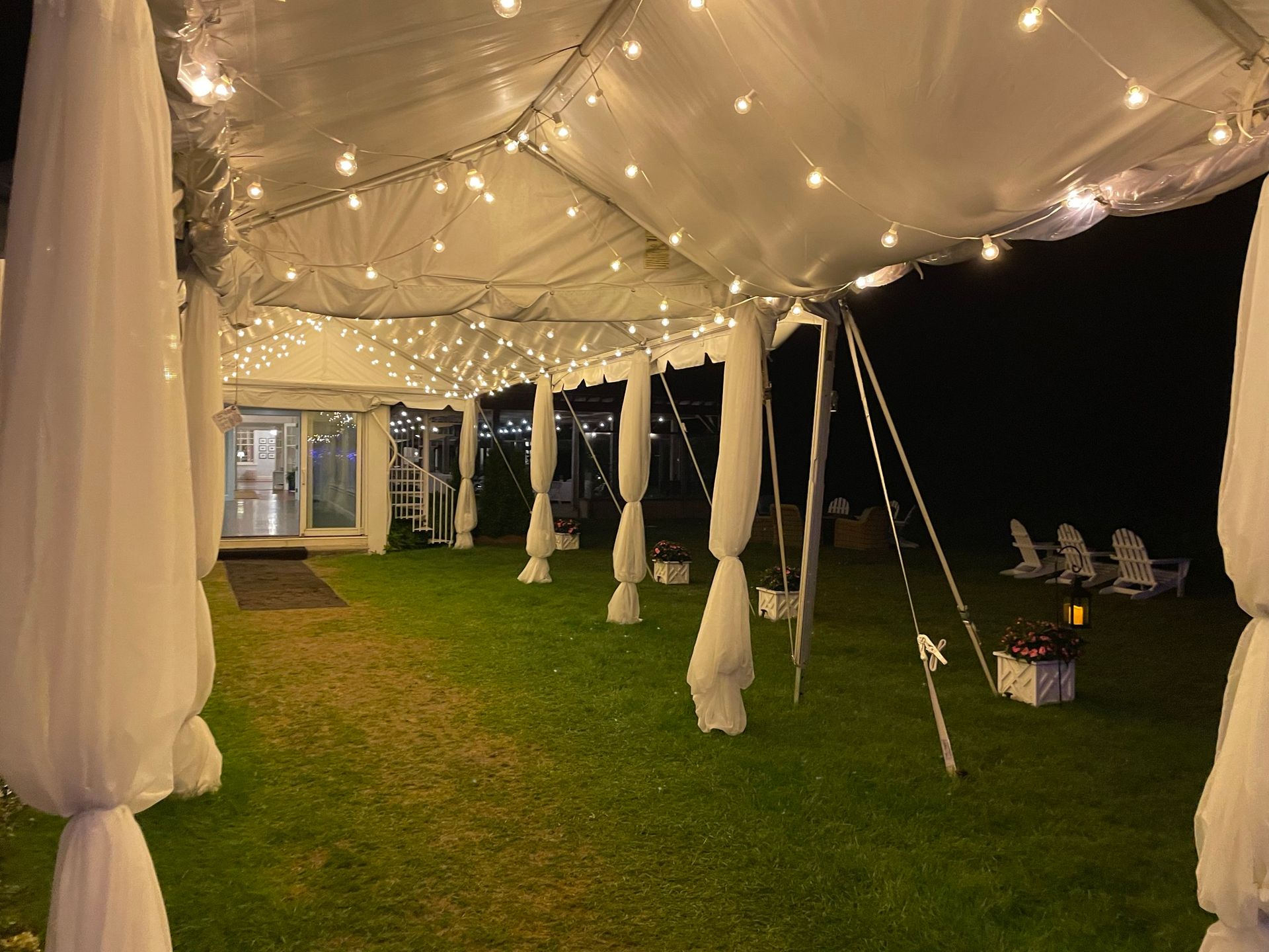 A lit-up tent decorated with string lights and white fabric draping on a grassy area, at night.