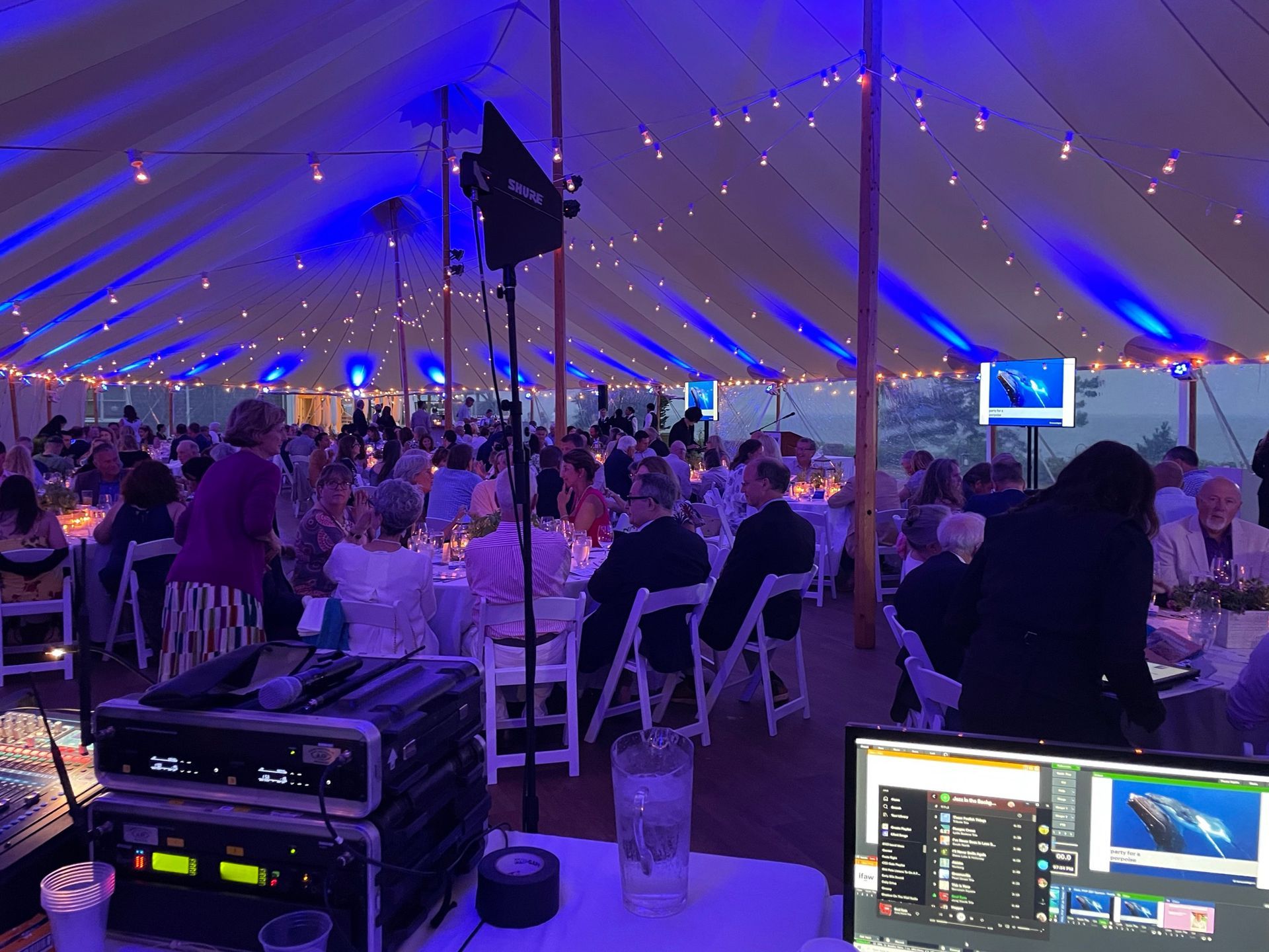 Large tent with tables set for a dinner, blue uplighting, people seated, DJ setup in foreground, monitors display a whale.