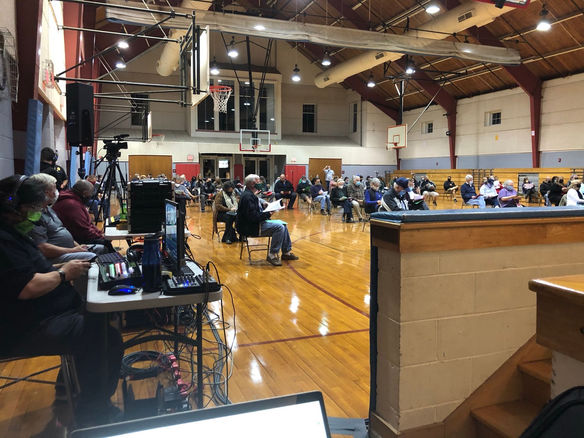 Gymnasium meeting: people seated, some wearing masks. Equipment on table; basketball hoops visible.