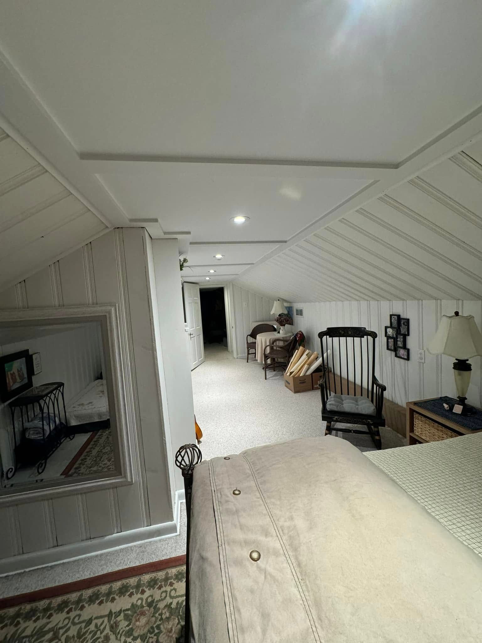 Loft area with white walls and ceiling, looking down into a long corridor with chairs and a bed.