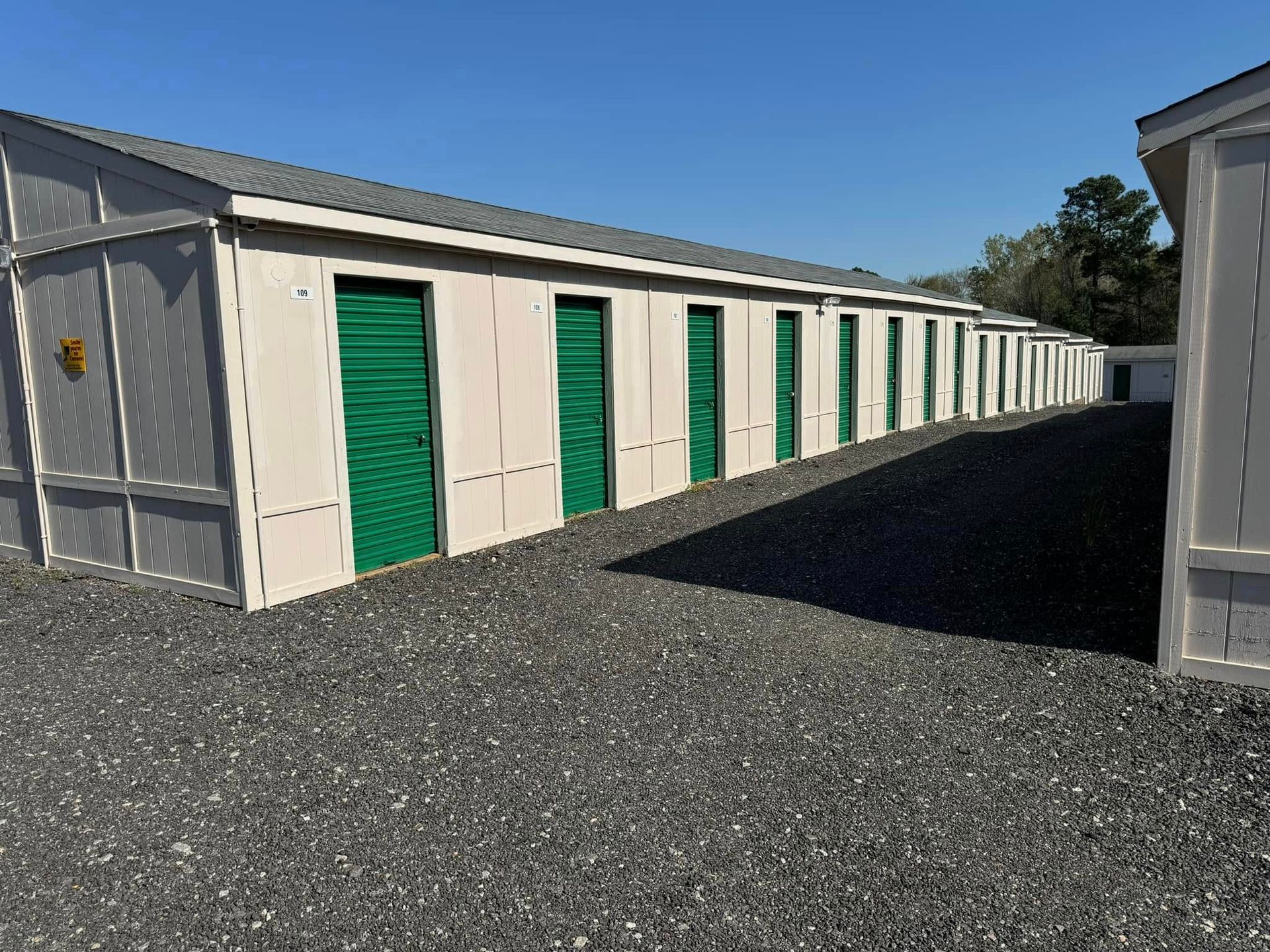 Storage units with green doors, arranged in a row, on a gravel lot under a blue sky.