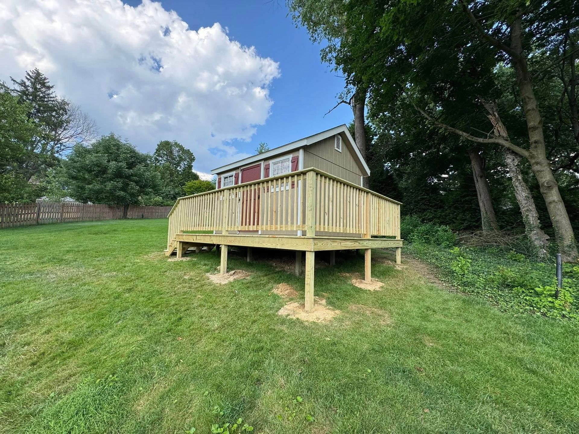 Wooden deck attached to small, wood-sided building, on a grassy hill, under a partly cloudy sky.