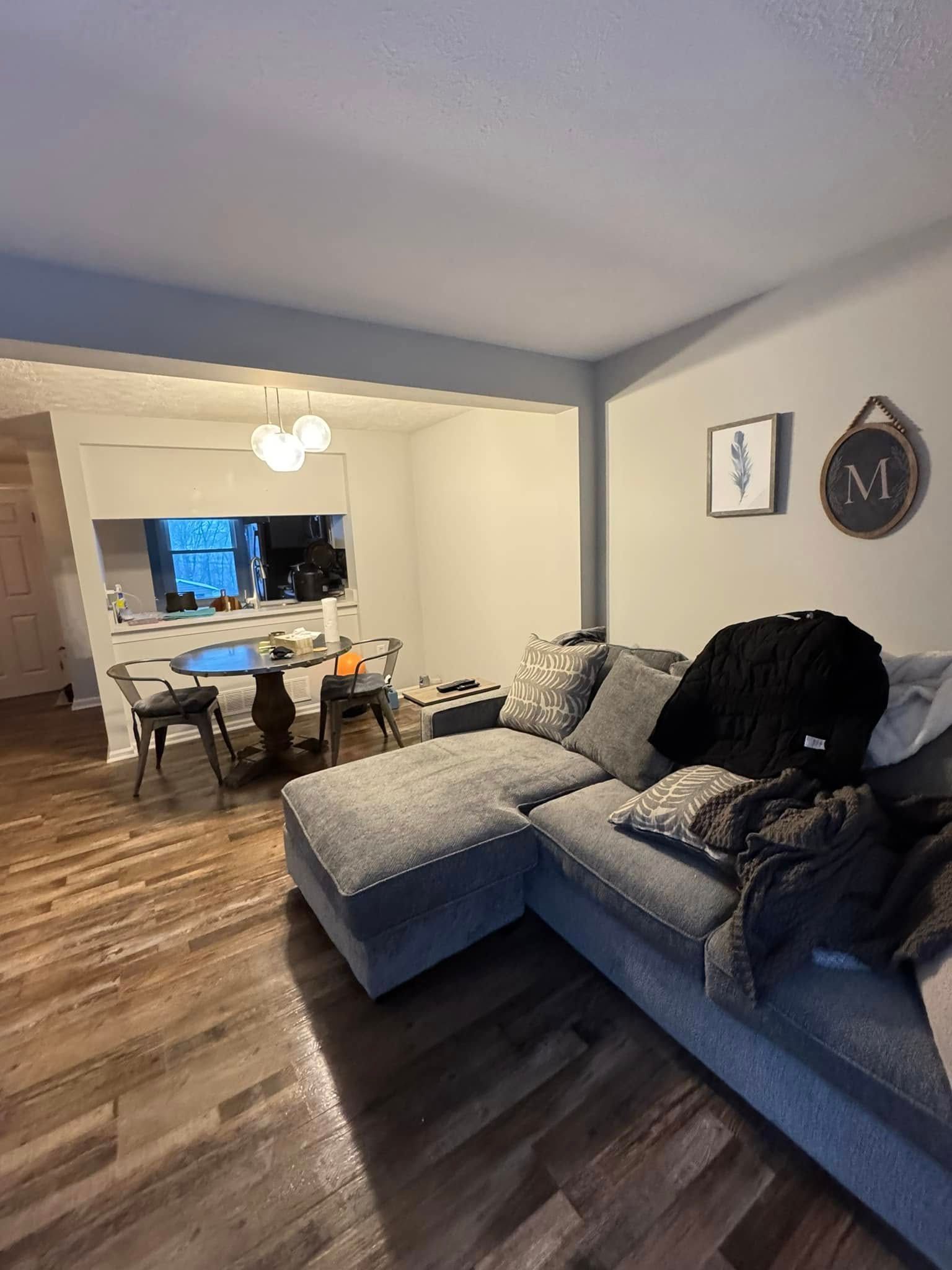 Gray sectional sofa in a living room with wooden floors, opening to a kitchen with a round table.