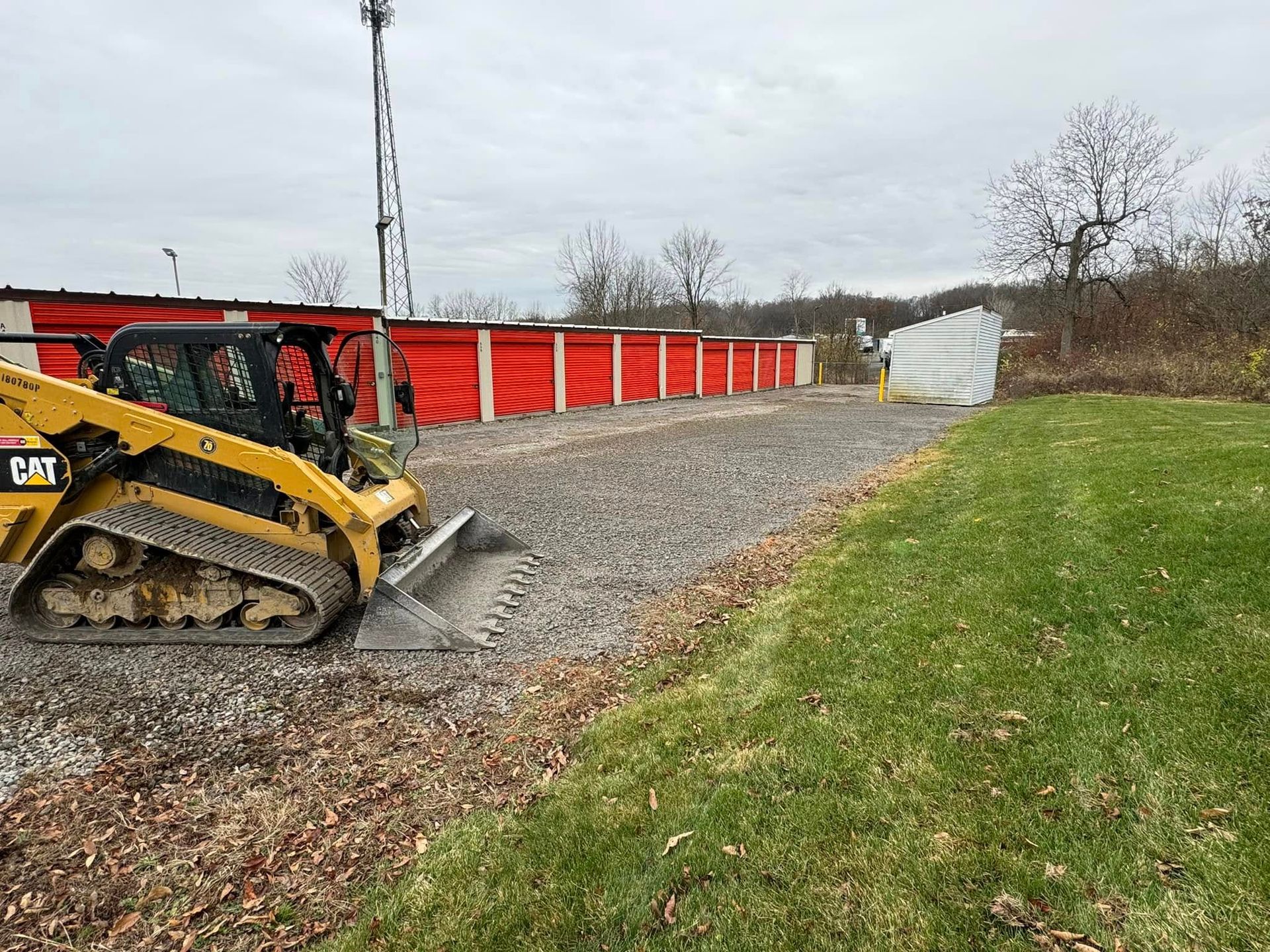 Yellow skid steer grading gravel in front of red barriers and a white building, on a cloudy day.