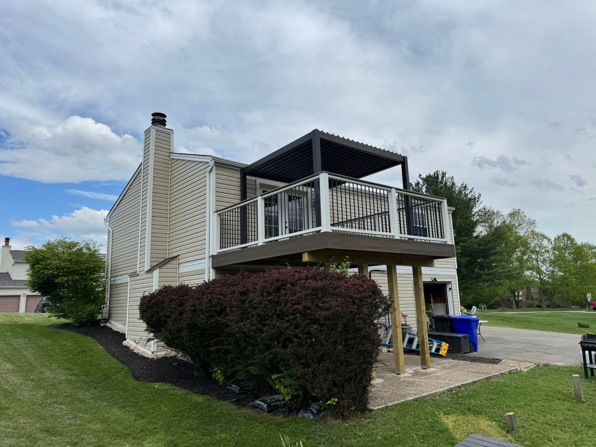 Two-story house with a wooden deck and black pergola on the upper level, a black-mulched garden, and a cloudy sky.