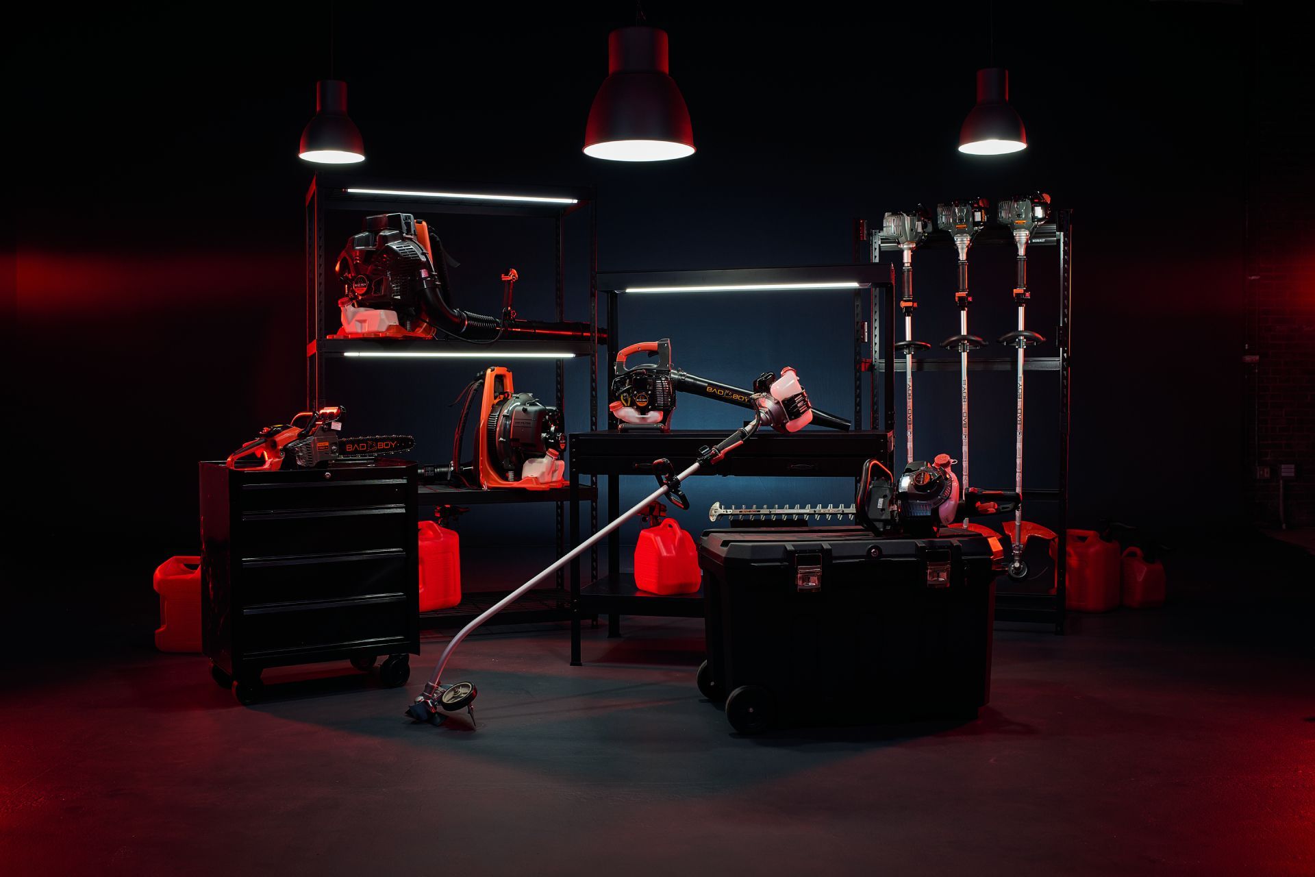 A dimly lit studio displays various orange and black outdoor power tools, including trimmers, blowers, and storage cases.
