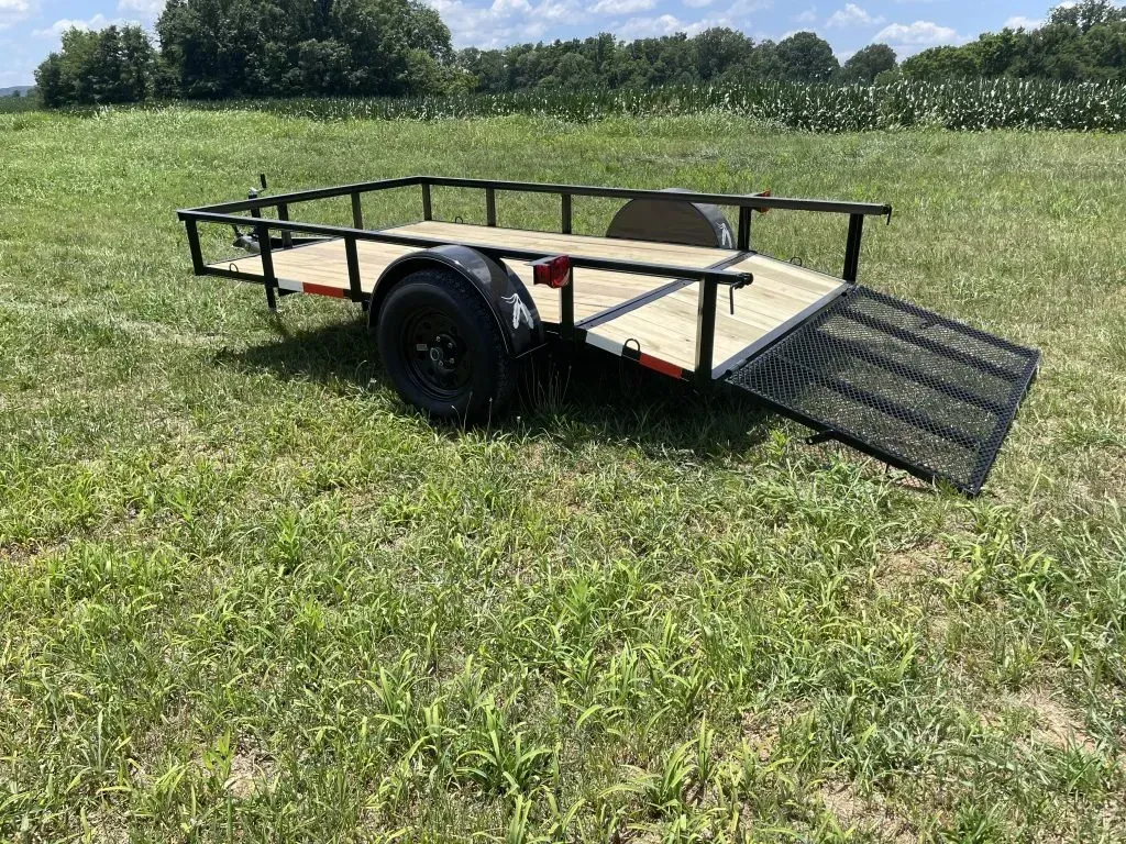 A black, single-axle utility trailer with a wooden floor and a mesh ramp sits in a grassy field.