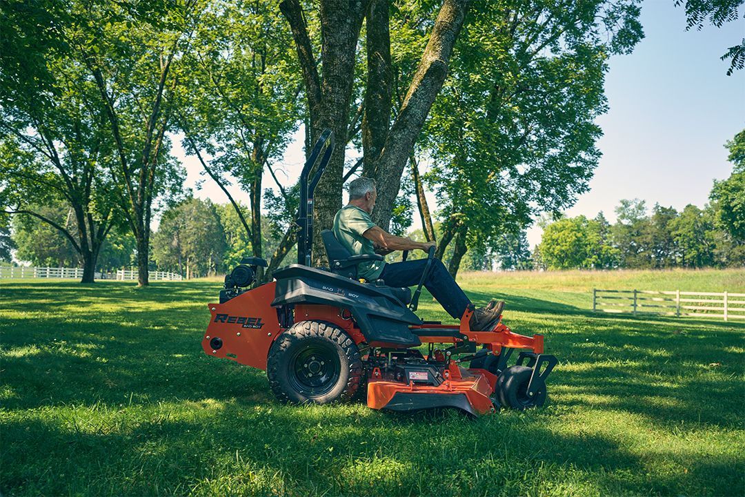 Person on a red zero-turn mower mowing grass near a tree in a sunny yard.