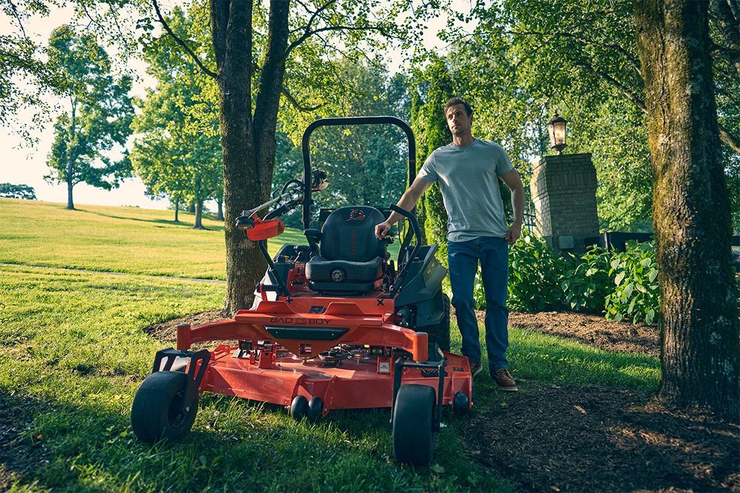 Man stands next to a red zero-turn mower in a grassy yard, trees in background.