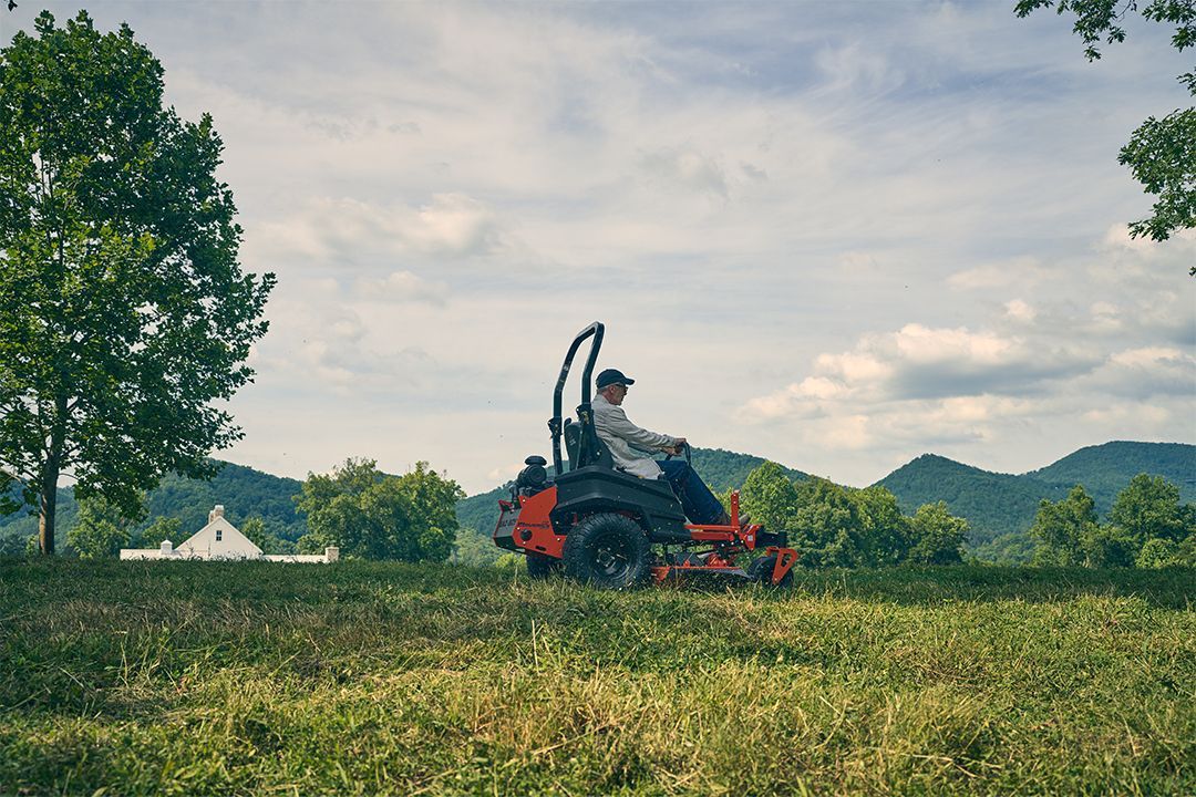 Person riding a red zero-turn mower in a field with mountains in the background.