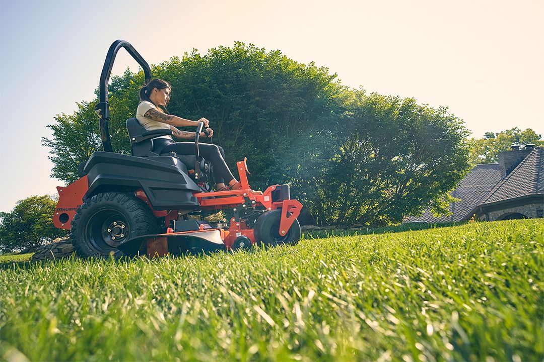 Person operating orange zero-turn mower on green grass near a large bush and house.
