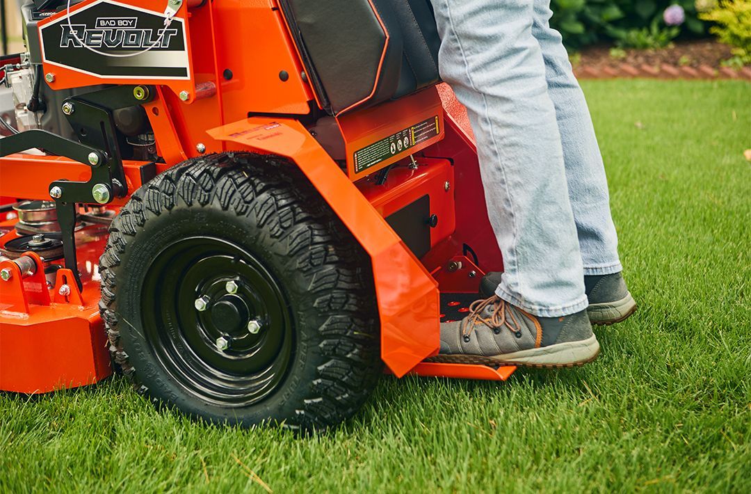 Person using a red and black lawn mower on a green lawn; legs and feet visible.