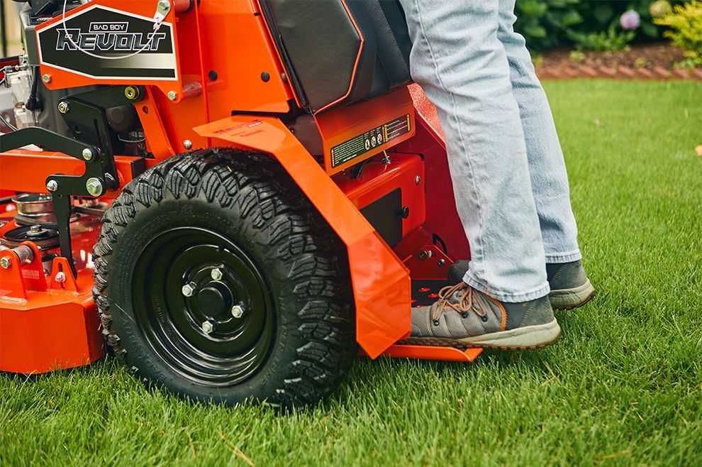 Person standing on the footrest of a red and black lawn mower.