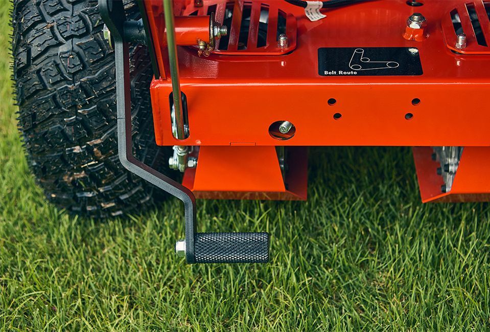 Orange lawn mower deck with black foot pedal, on green grass.