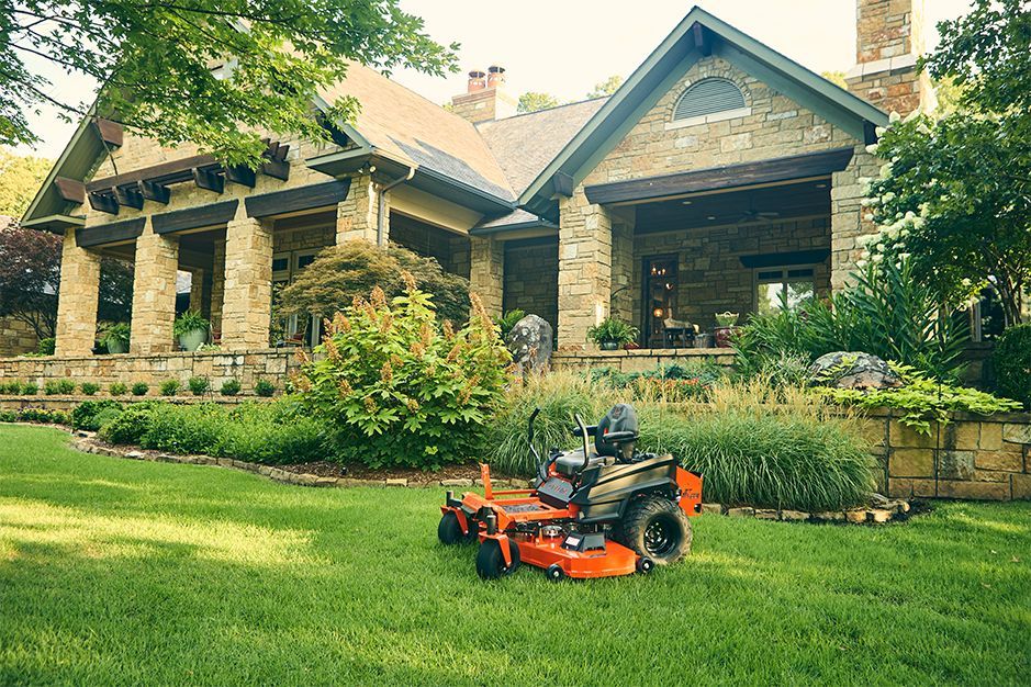 Orange riding lawnmower on a green lawn in front of a stone house with porch and landscaping.