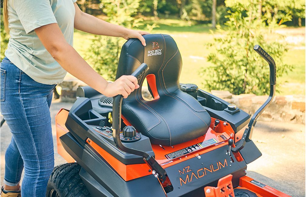 Woman seated on an orange and black zero-turn lawnmower, grasping the armrests, in an outdoor setting.