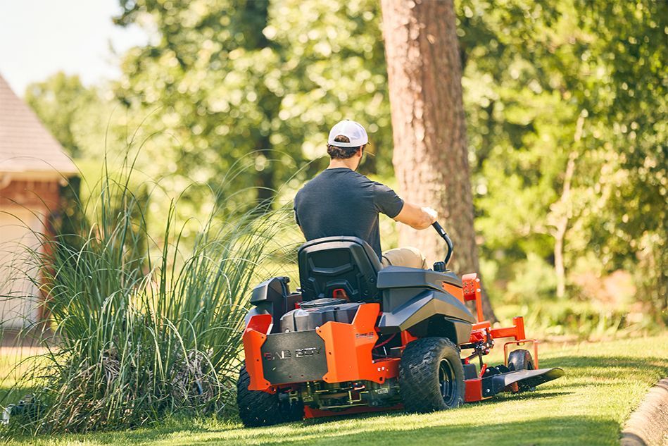 A person on a red zero-turn mower cutting grass near a tree in a yard.