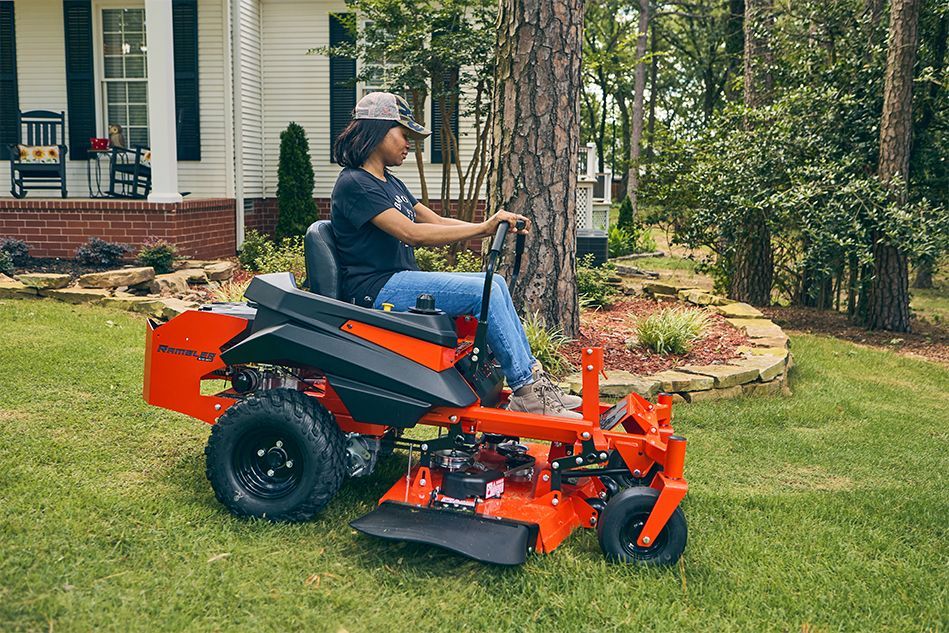 Person driving an orange riding lawnmower on a green lawn near a house and trees.