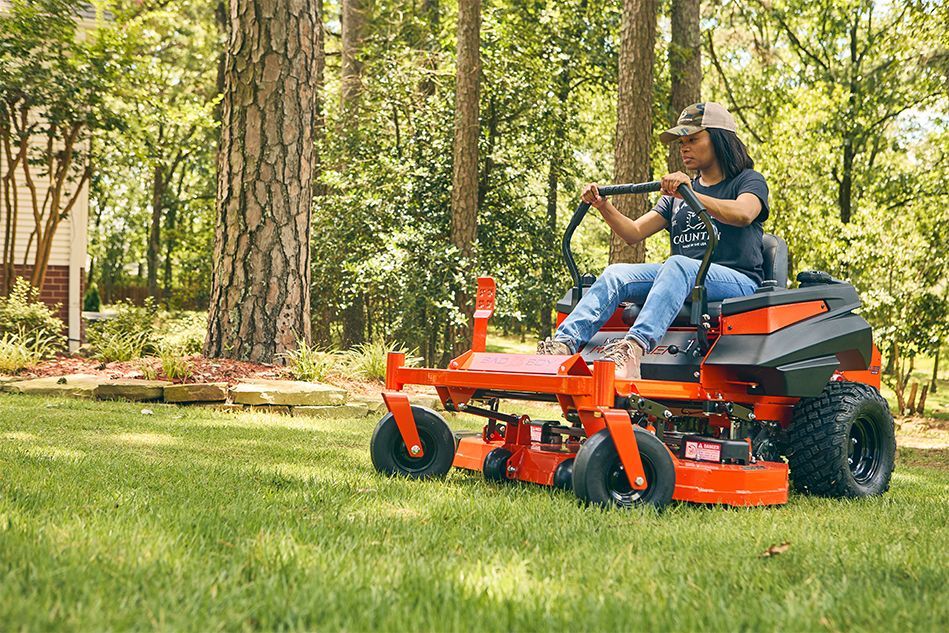 Person driving an orange zero-turn lawnmower on grass in a wooded yard.