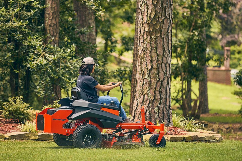 Person on a bright orange riding lawnmower mowing a green lawn near trees.