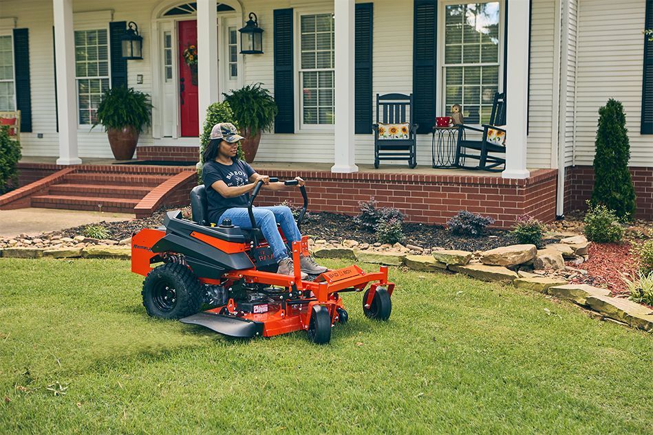 Person on orange riding lawnmower mowing a green lawn in front of a white house with red door.