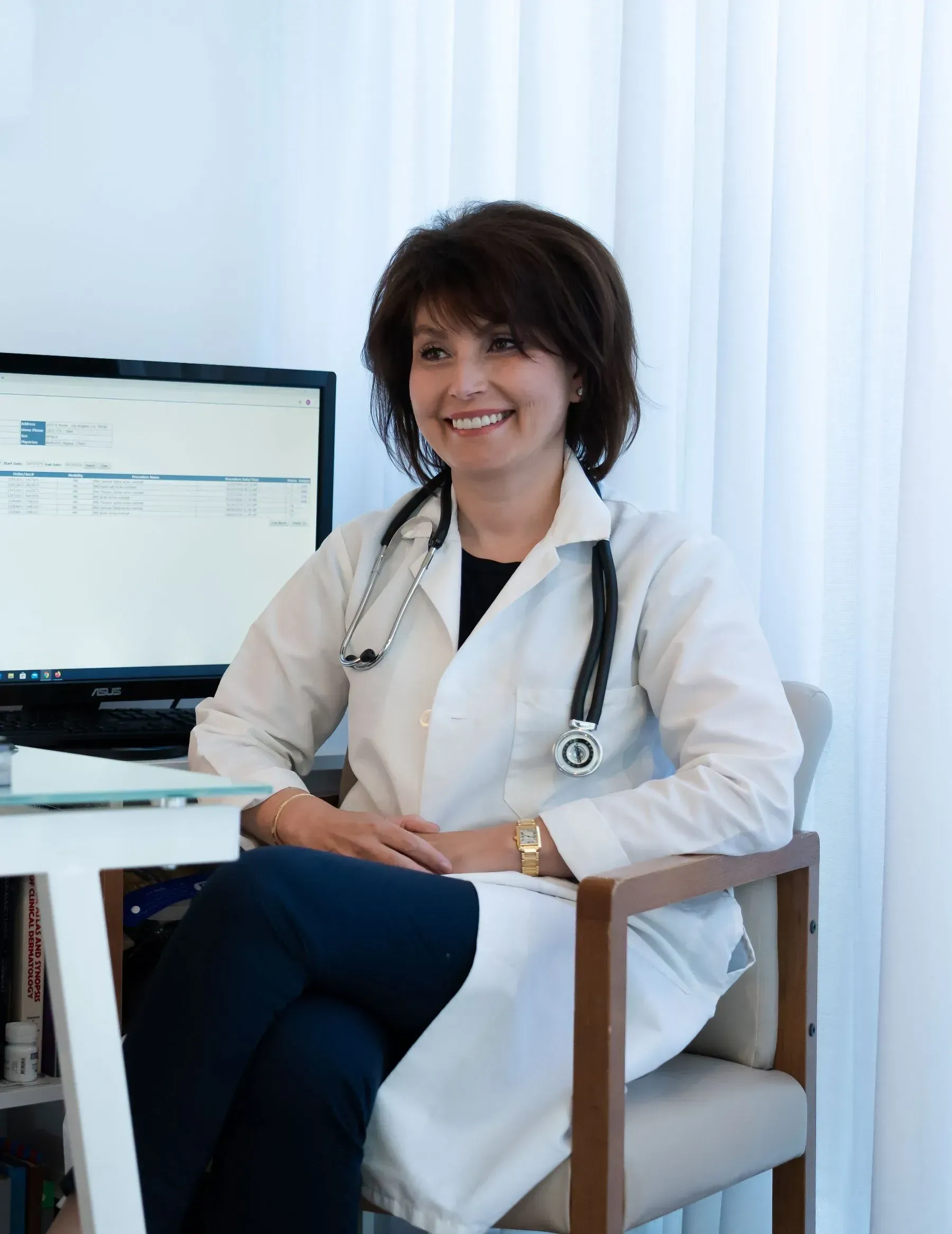 Doctor in white coat smiling at a desk beside a computer in a bright office