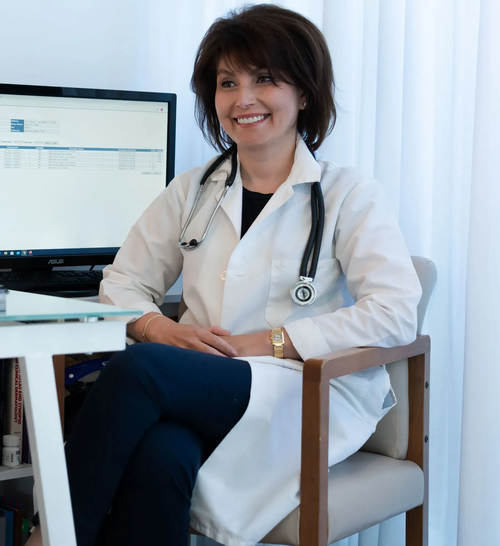 Smiling doctor in a white coat sits beside a computer in a bright office