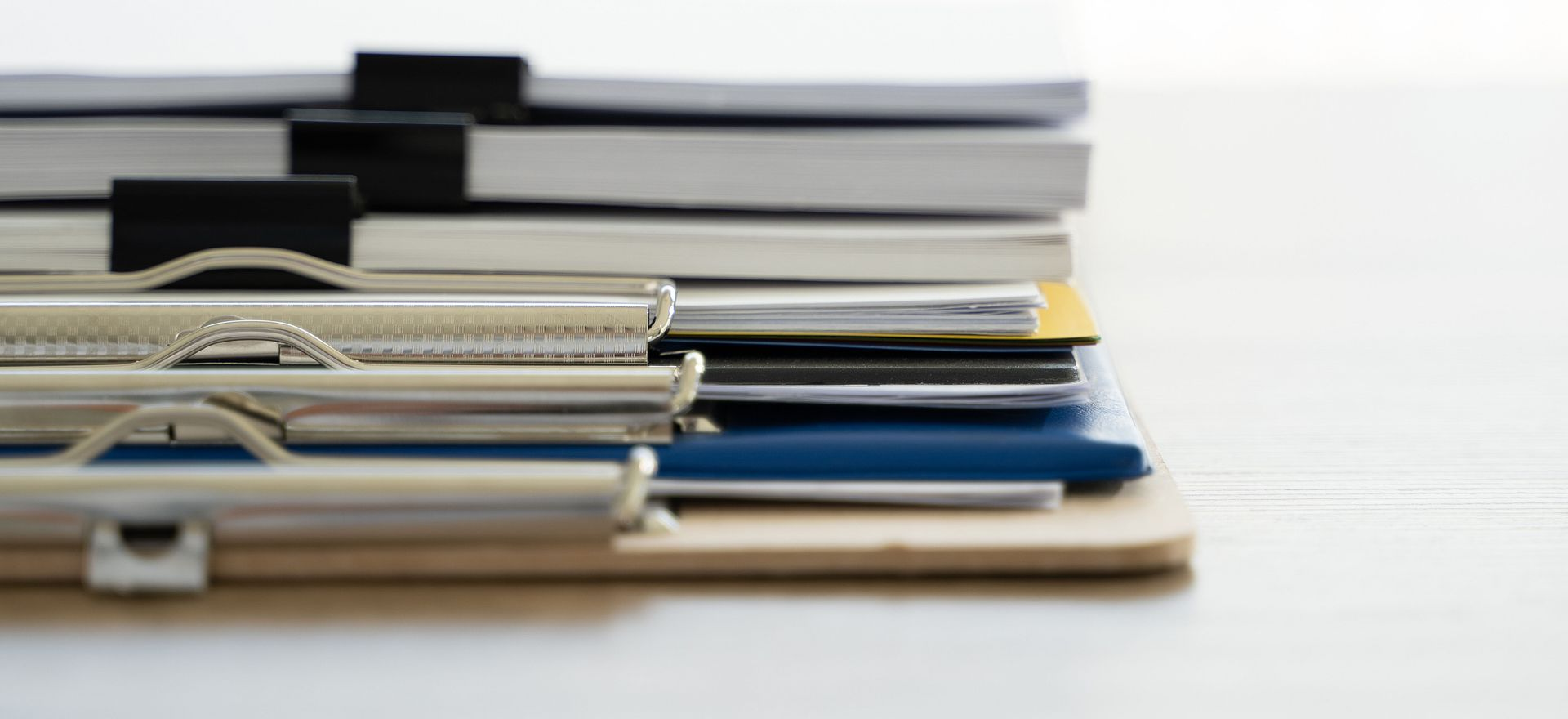 A stack of papers with binder clips, folders, and a clipboard on a wooden surface.