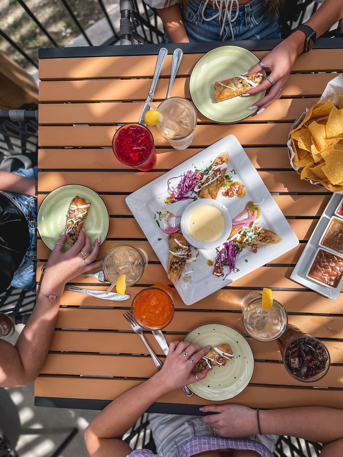 Overhead view of a wooden table with food and drinks. People are reaching for tacos, chips, and cocktails outdoors.