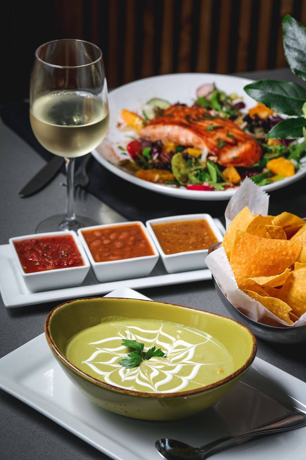 Bowl of soup, chips, salsa, salmon salad, and white wine on a table.