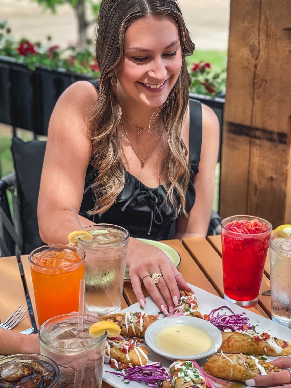Woman smiling, seated at outdoor table with food and drinks.