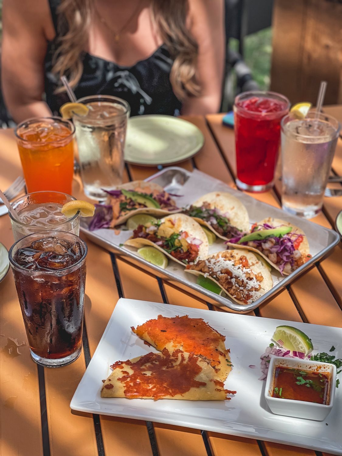 Table with tacos, drinks, and a side dish. Outdoor setting with sunlight.