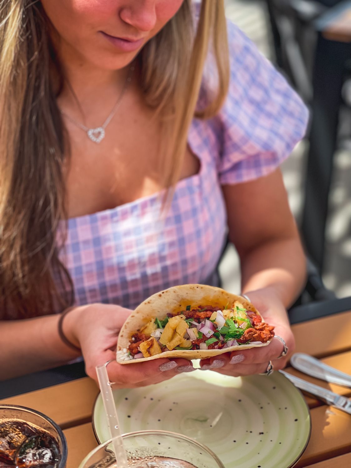 Woman holding a taco, seated outdoors. The taco has fillings of various colors. She wears a pink plaid top.